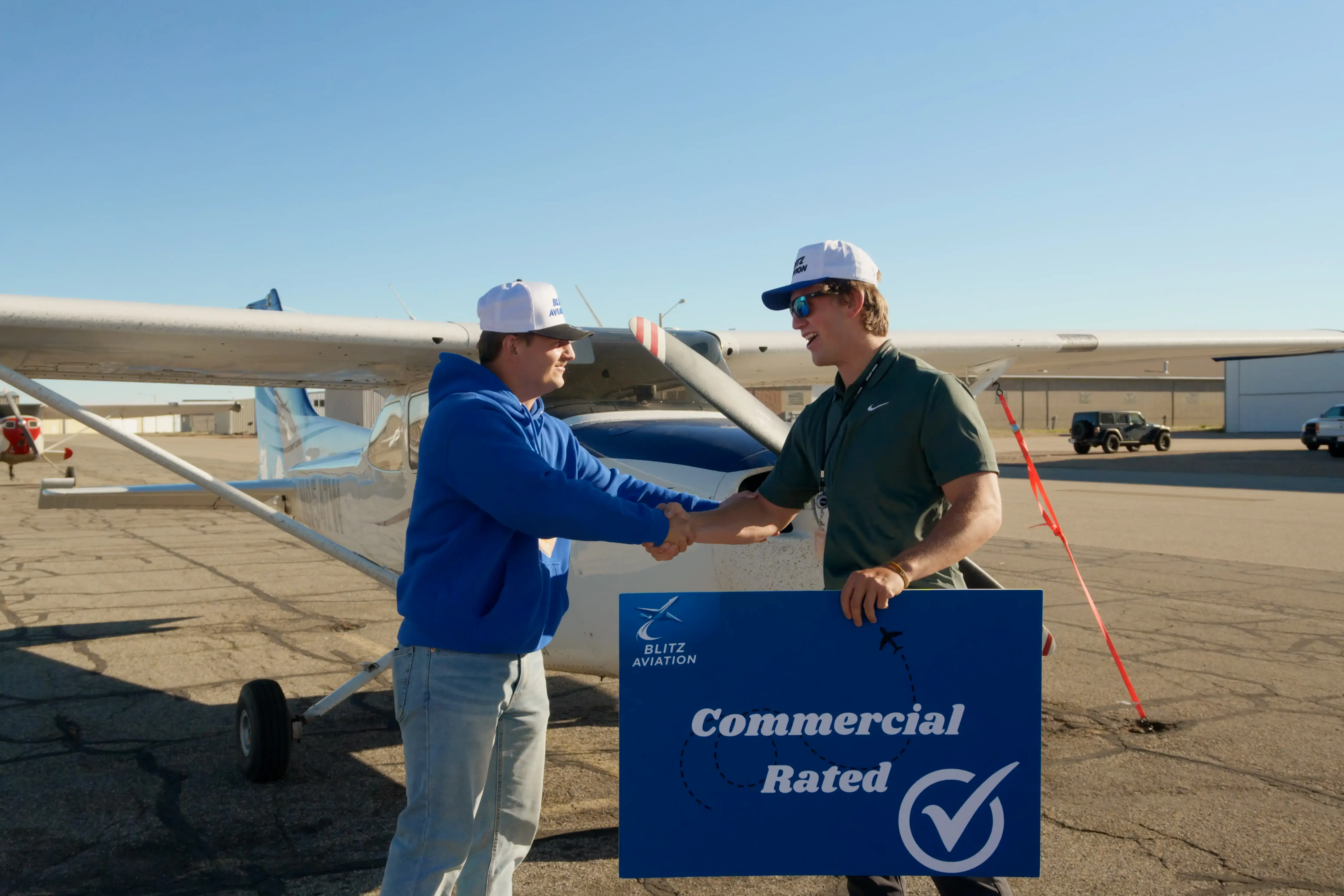 Student and CFI holding hands in front of an airplane while holding a poster with "Commercial Rated" on it