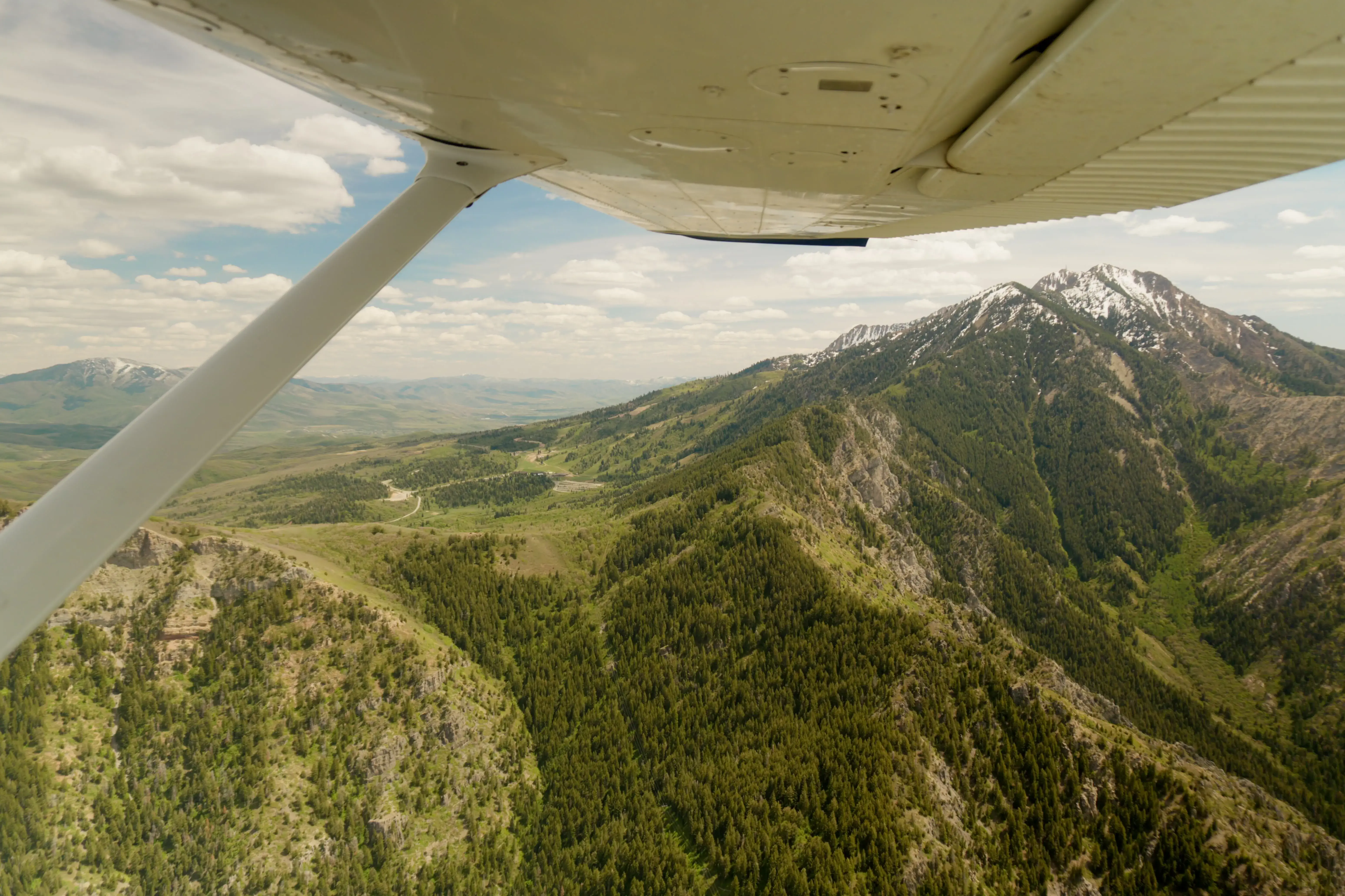 View from under the wing of a Cessna over forested mountains with snow-capped peaks and scattered clouds